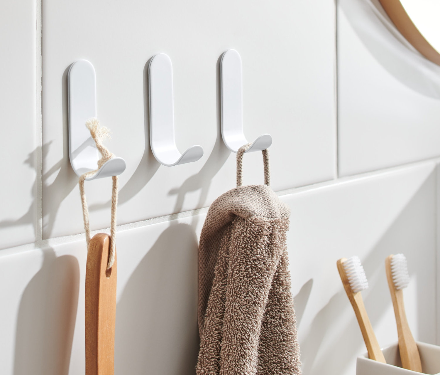 A fehér csempén három öntapadós akasztó lóg, rajtuk egy kefe és egy törölköző.