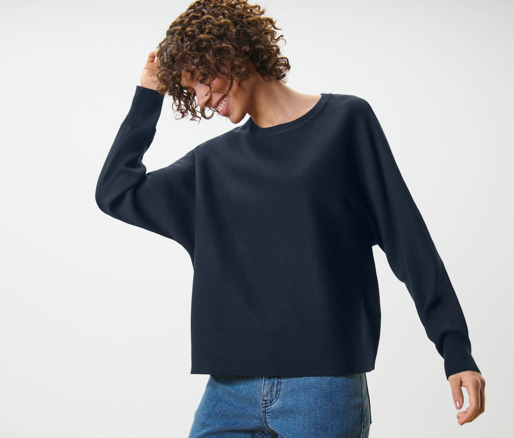 Göndör hajú nő mosolyog és megérinti a haját, sötétkék finomkötésű denevérujjú pulóvert és kék farmert visel.