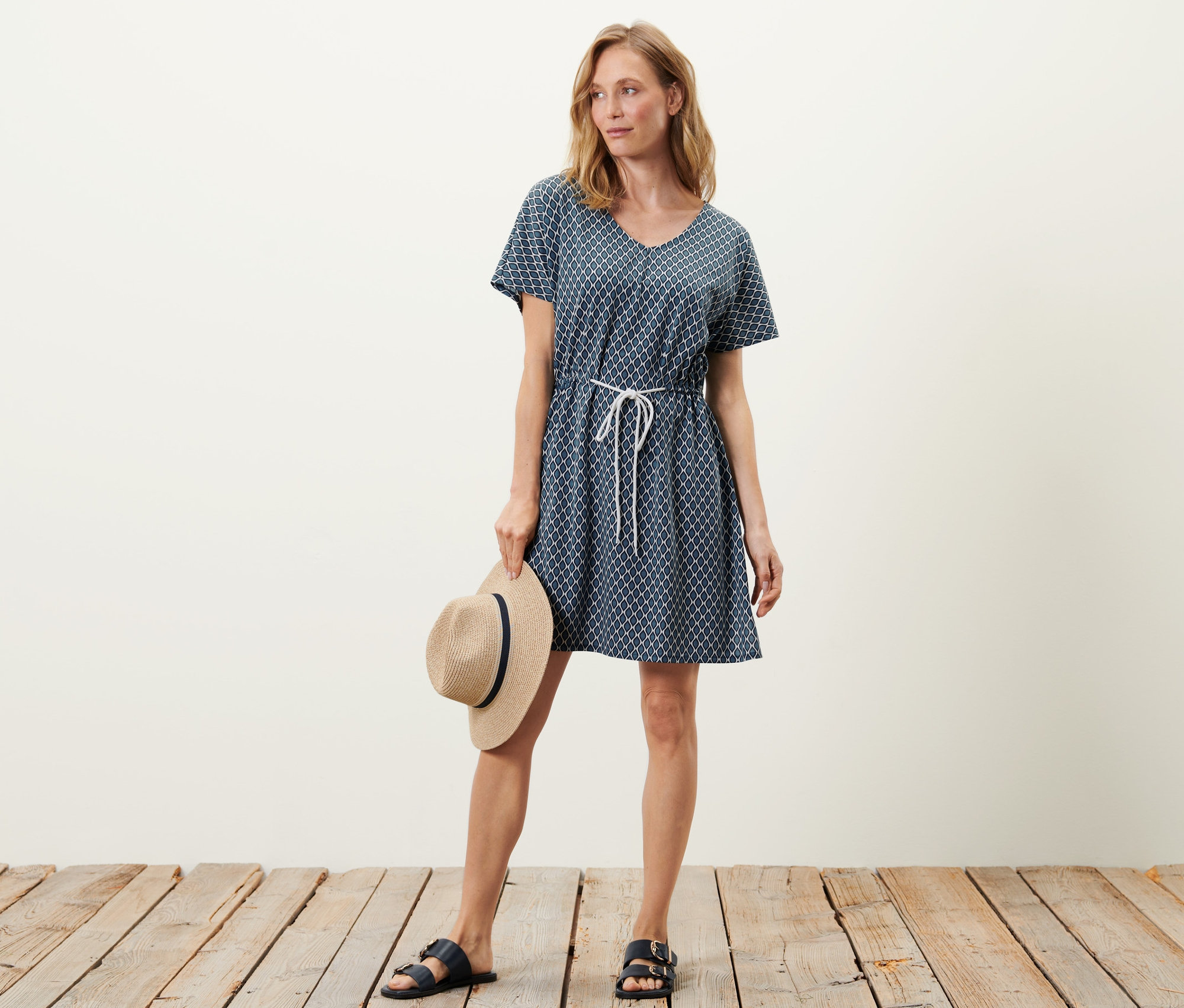 Nő strandtunika ruhában pózol és egy kalapot tart.