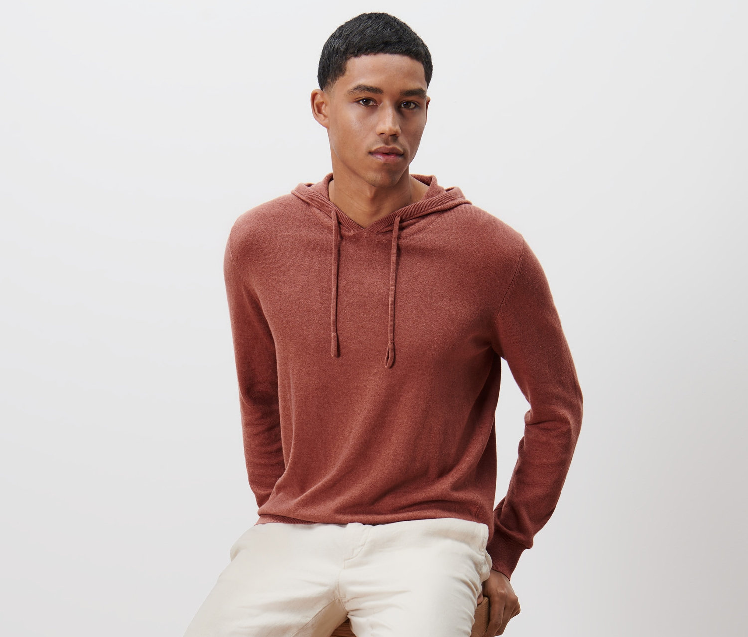 Egy férfi ül egy fa zsámolyon, és kötött kapucnis felsőt visel.