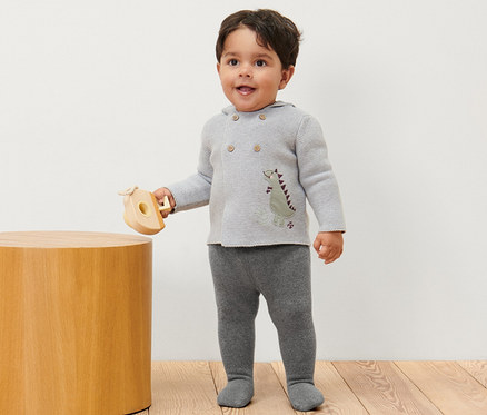 Egy kisfiú áll és egy fa játékot tart, melírozott termo harisnyanadrágot visel.