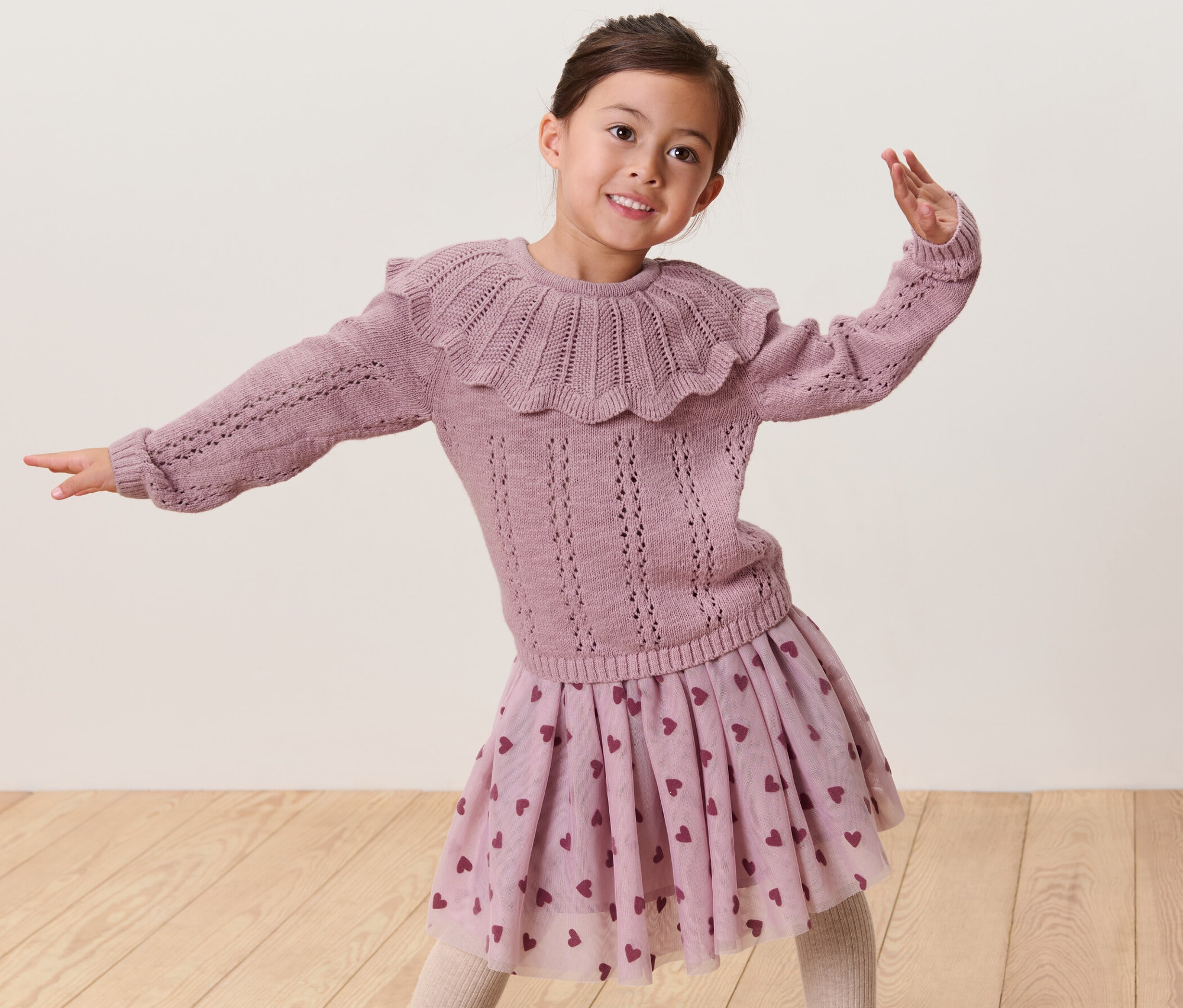 Mosolygó lány pózol egy kislány rózsaszín kötött pulóverben és egy szívecskés szoknyában.