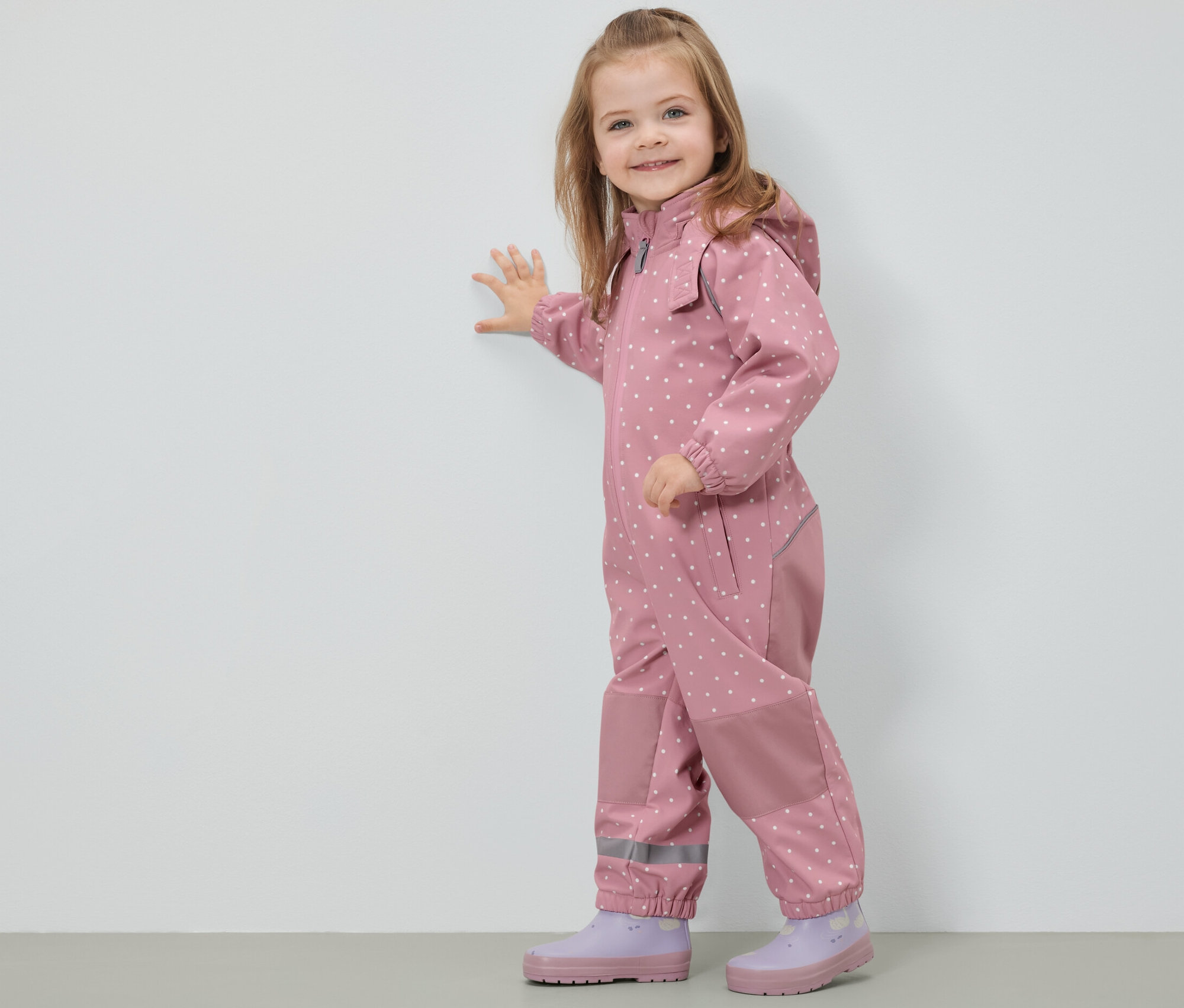 Egy mosolygó kislány pöttyös gyermek softshell kezeslábasban áll a falnál.