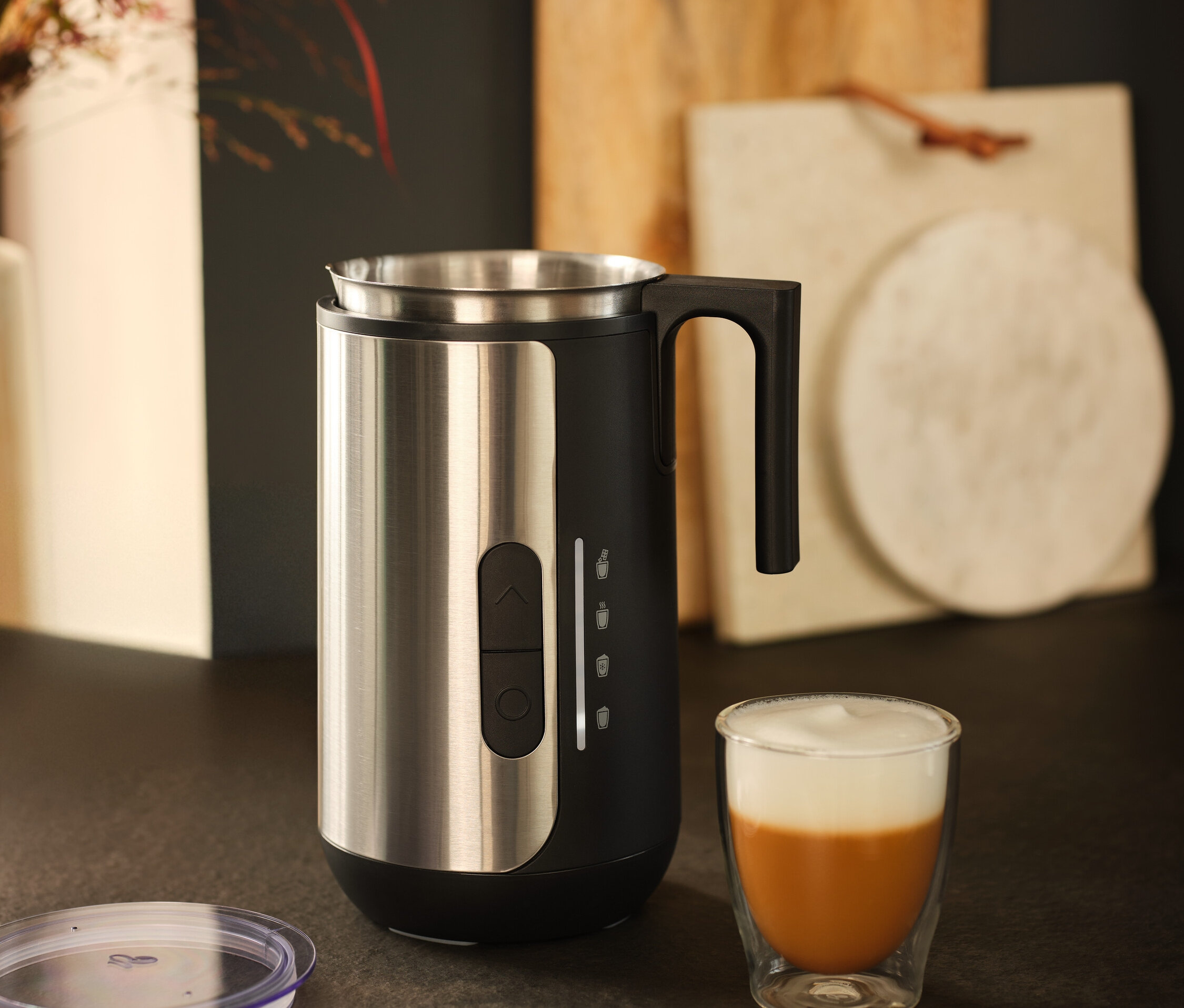Egy rozsdamentes acélból készült tejhabosító áll egy pohár tejeskávé mellett.