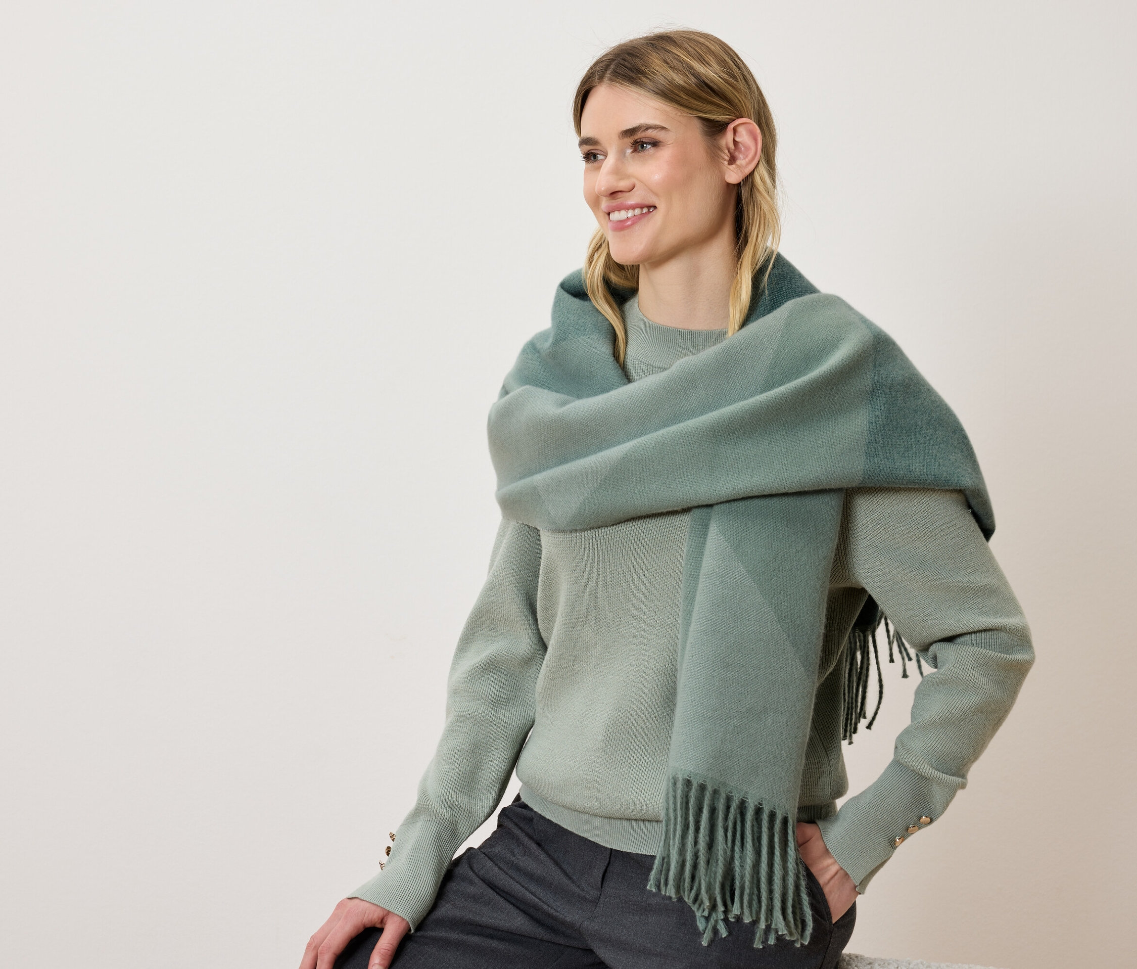 Egy mosolygó nő zöld pulóvert és kockás, rojtokkal ellátott sálat visel.