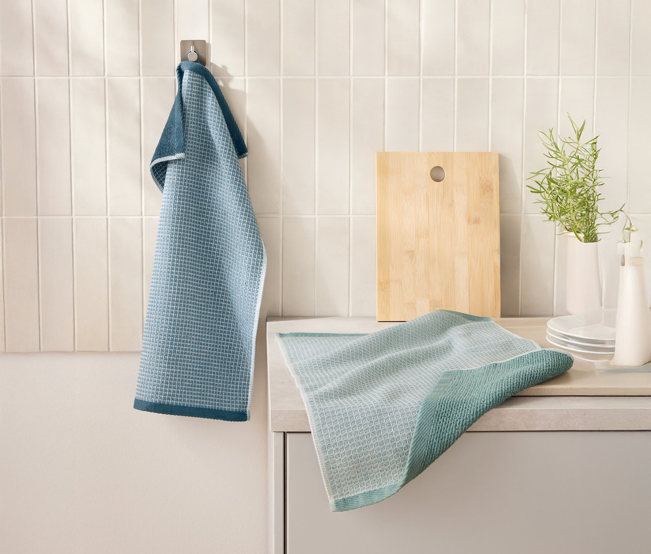 Egy kék 2 Waffelpiqué-Küchenhandtücher lóg egy akasztón egy vágódeszka mellett. Egy másik konyharuha egy konyhapulton fekszik.