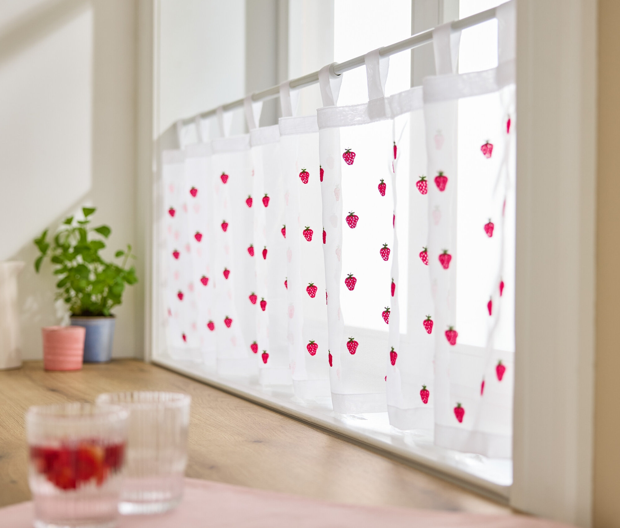 Egy hímzett félfüggöny látható az ablakon, eper mintával.