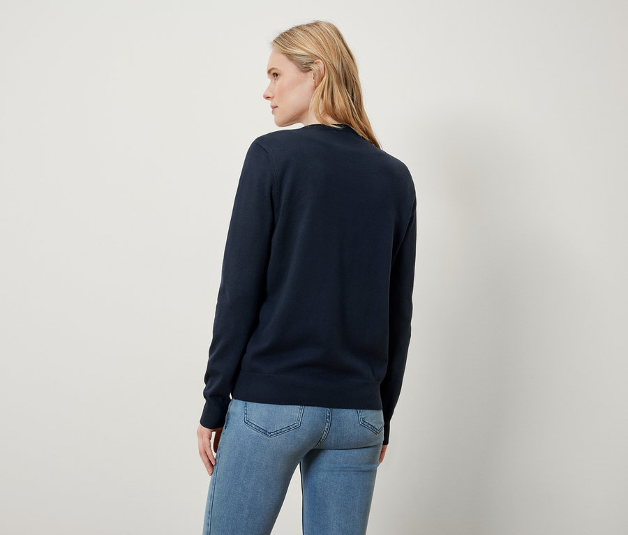 A szőke nő háttal áll a kamerának, sötétkék pulóvert és farmert visel.