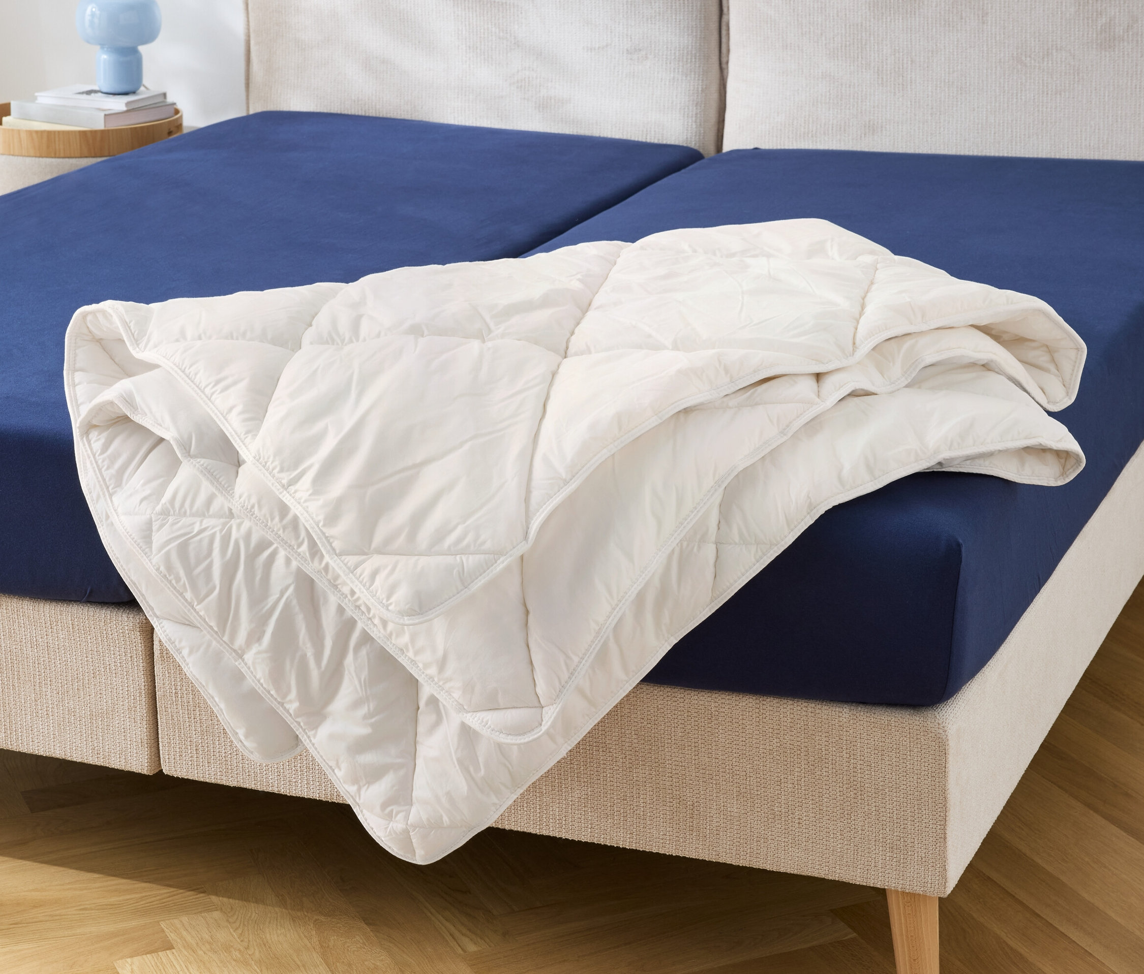 Egy kék lepedővel borított ágyon egy fehér, steppelt takaró fekszik.