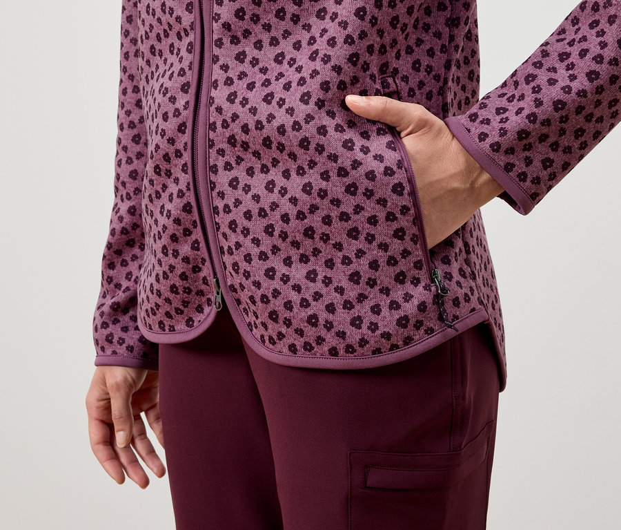 Nő kezét a lila mintás kabát zsebében tartja, lila nadrággal viselve.