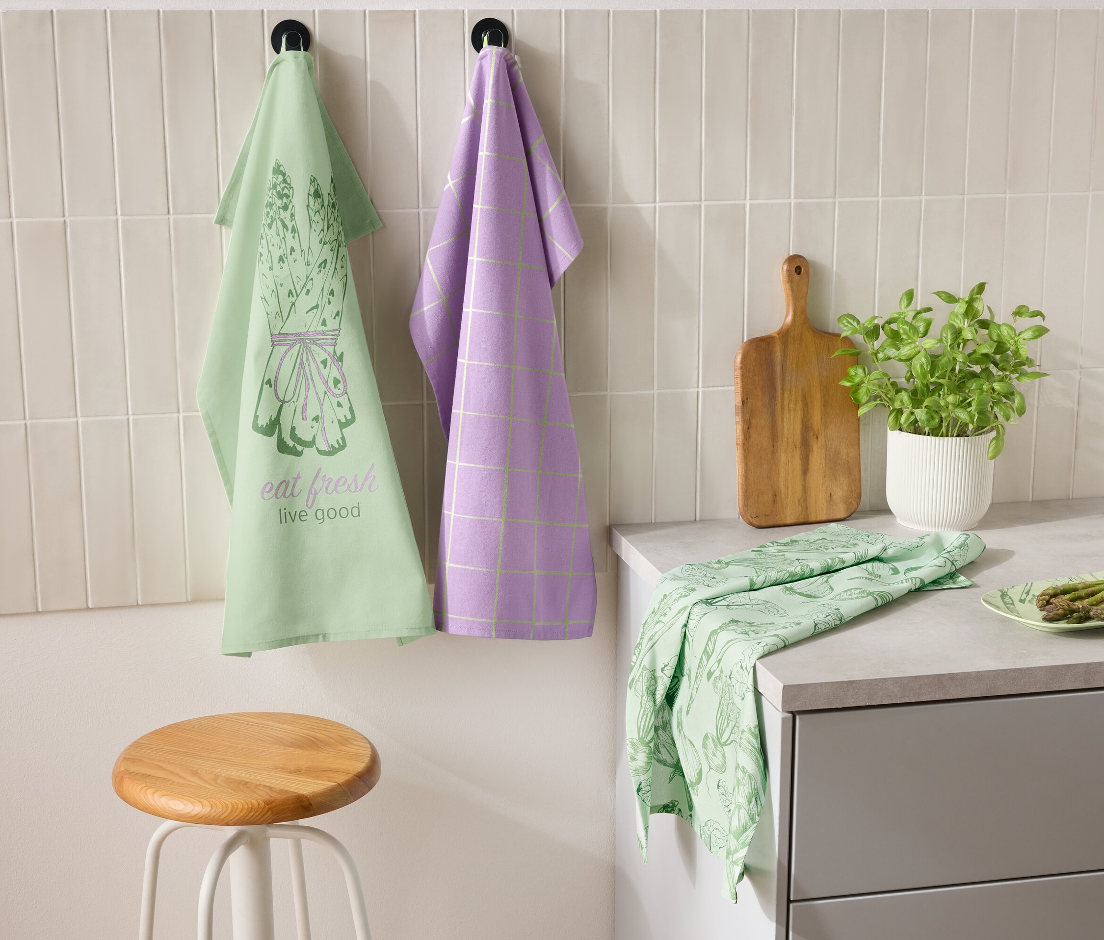 Két konyharuha lóg egymás mellett a falon, az egyik zöld spárga mintával és "eat fresh live good" felirattal, a másik lila kockás mintával. A pulton egy zöld konyharuha és egy tányér spárga fekszik. Mellette egy vágódeszka és egy fűszernövényes cserép áll.