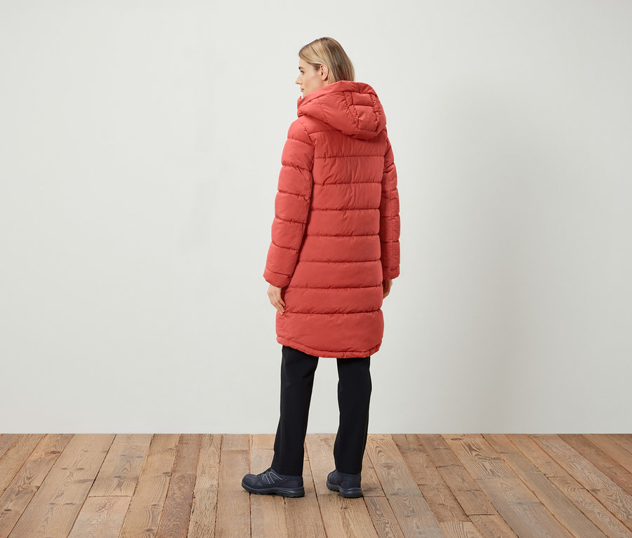 Egy nő áll háttal, piros, steppelt, kapucnis kabátot, fekete nadrágot és sötét cipőt visel.