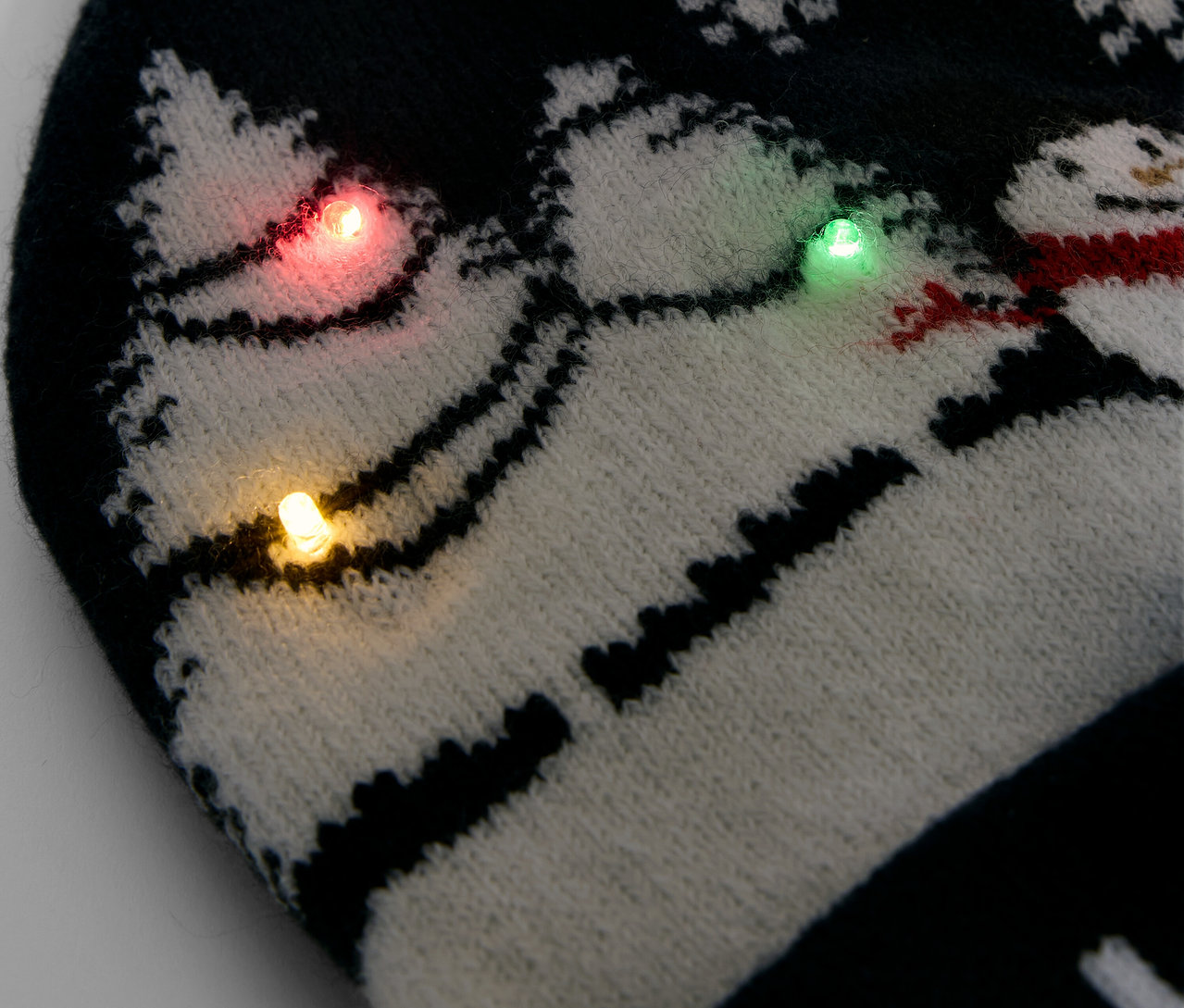 Egy fekete-fehér kötött sapka részlete hópihékkel és hóemberekkel, színes fényekkel díszítve.