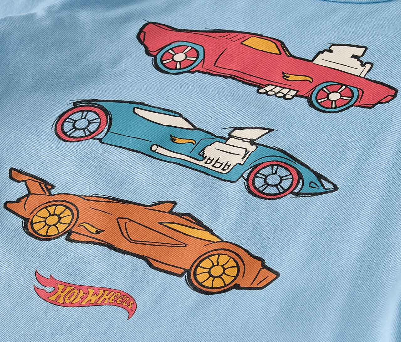 Egy világoskék pólón három piros, kék és narancssárga rajzolt Hot Wheels autó látható.
