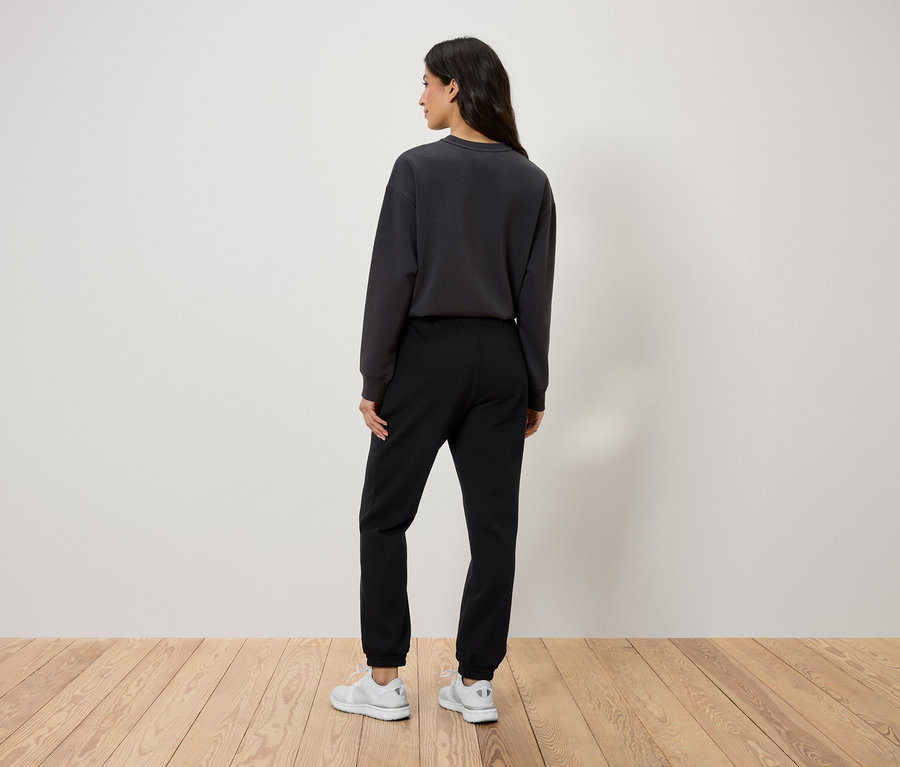 Hátulról látható egy nő fekete joggingalsóban, szürke pulóverben és fehér cipőben.