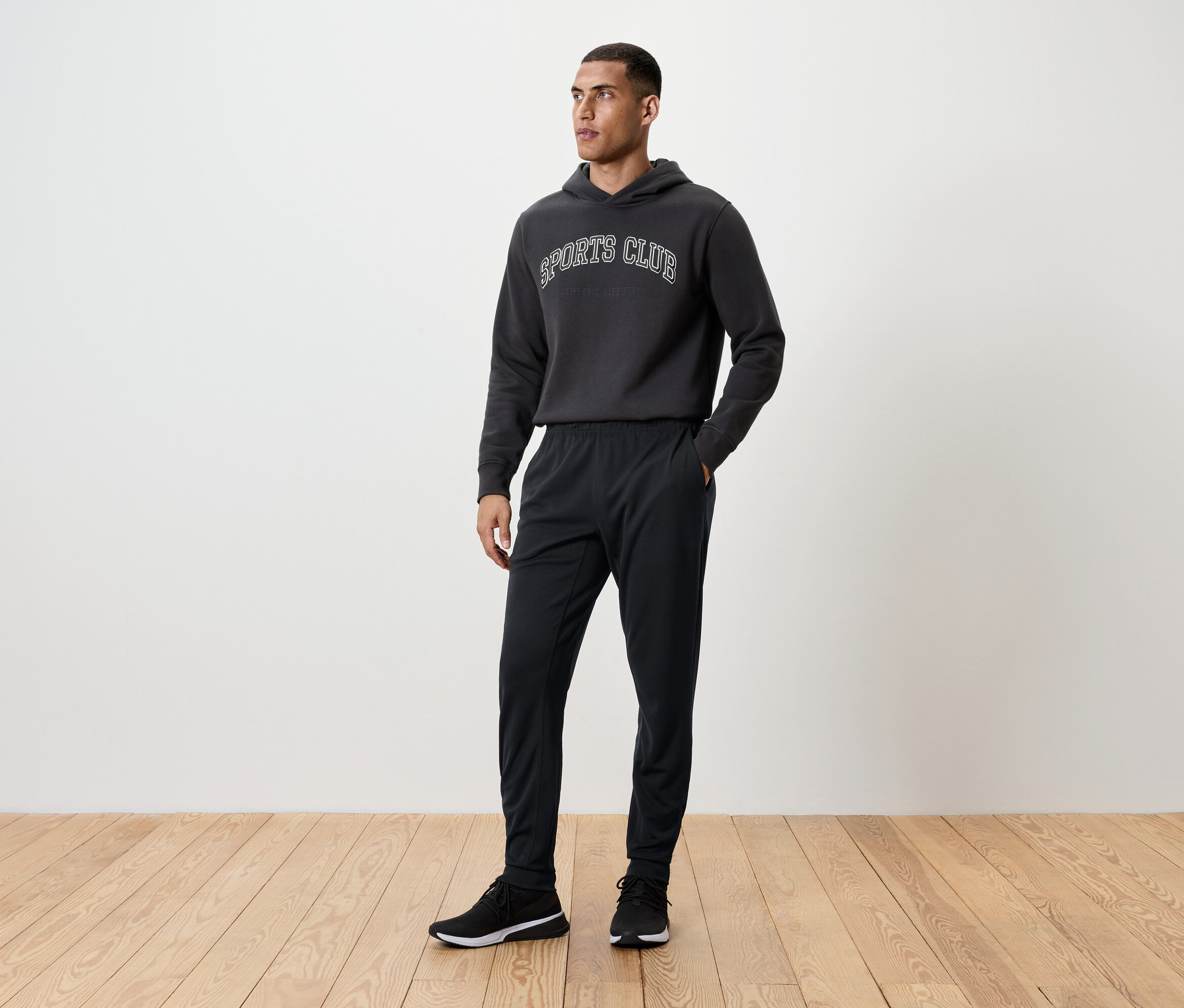 Férfi áll a fapadlón, fekete edzőnadrágot, kapucnis pulóvert és tornacipőt visel.