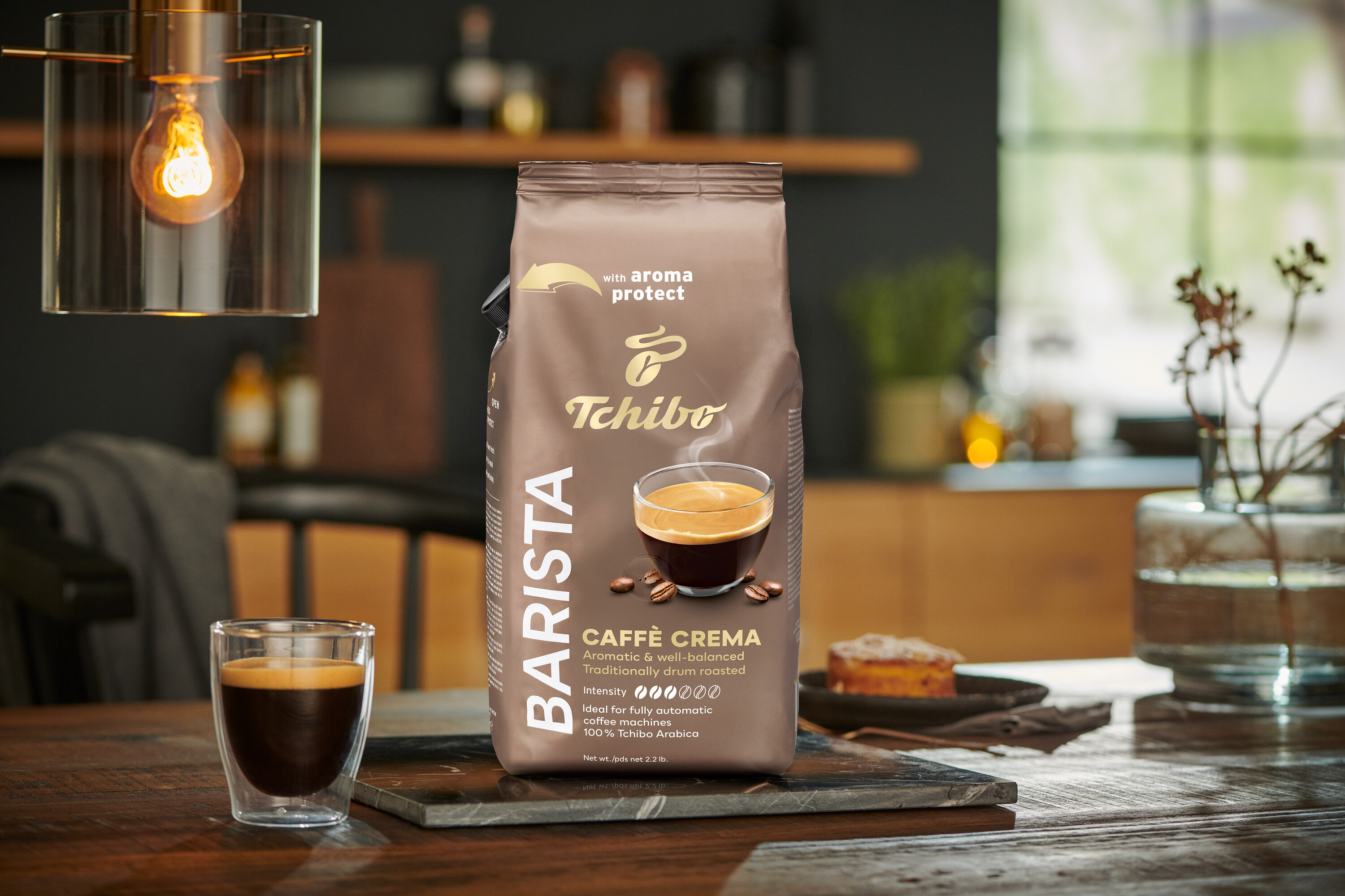 Egy csomag BARISTA Caffè Crema - 1x1 kg szemes kávé egy asztalon áll egy pohár kávé mellett.