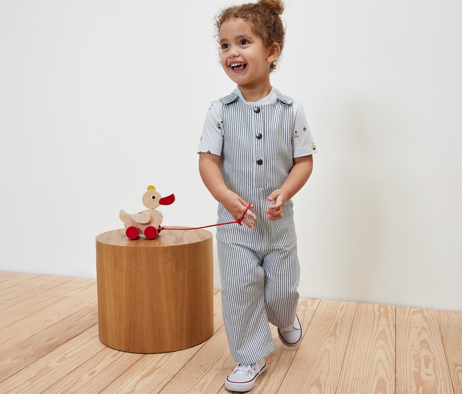 Mosolygó gyermek húz egy kerekes fa kacsa játékot, és szövet kantáros nadrágot visel.