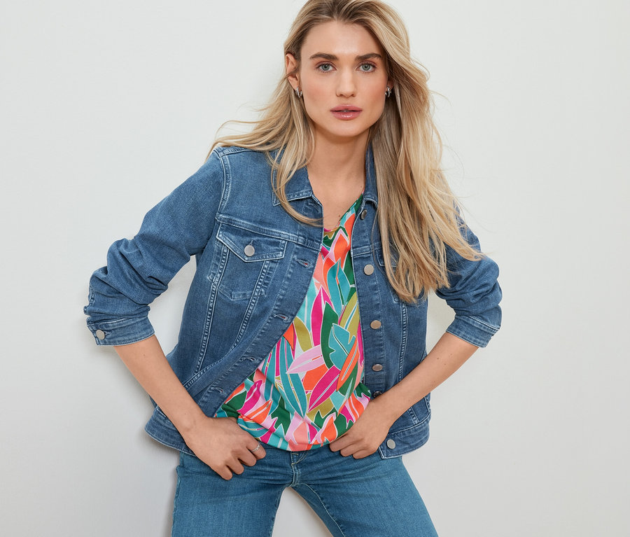 Nő pózol farmerdzsekiben, nyomott mintás pólóblúzban és egyenes szárú farmernadrágban.