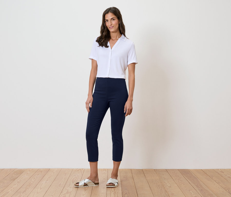 Nő áll a fal előtt, fehér inget, kék nadrágot és arany szandált visel.