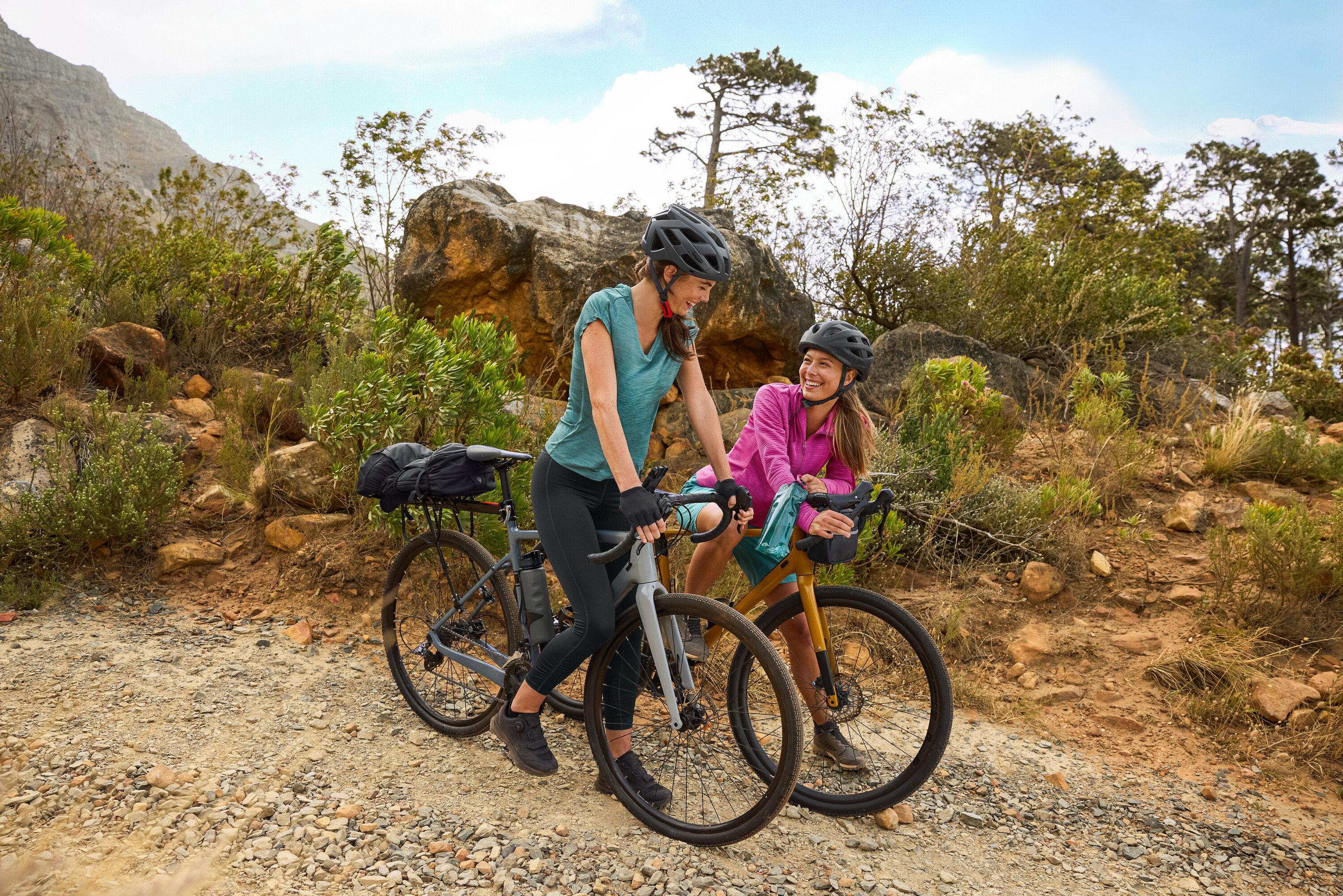Két nő sisakban áll a kerékpárjaival egy dombos vidéken, egy kavicsos úton. Egy kulacs van a kerékpárra rögzítve.