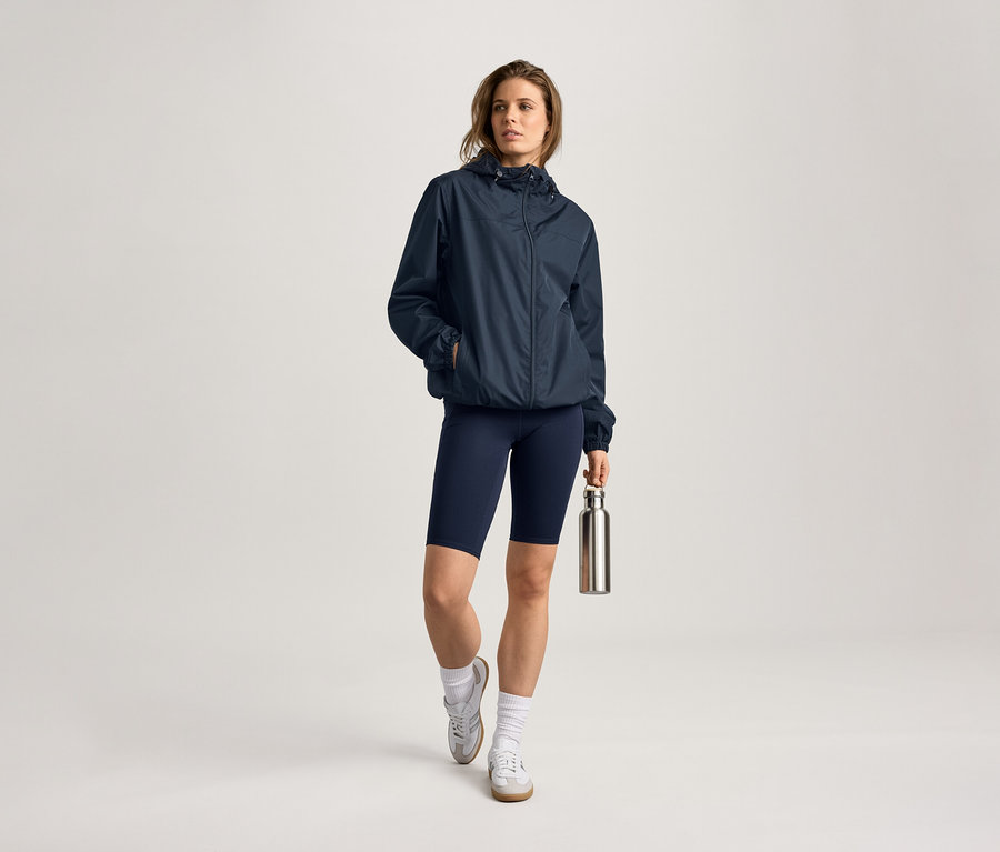 Nő esőkabátban, kerékpáros nadrágban és kulaccsal. Zoknit és tornacipőt visel.