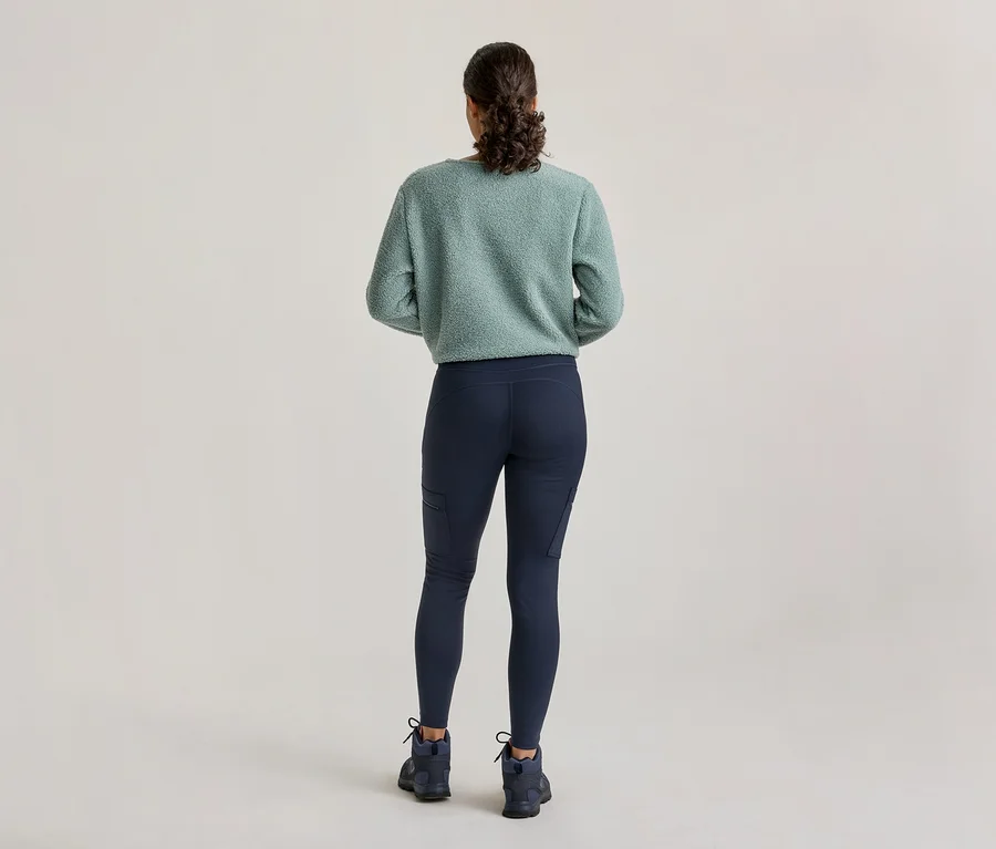 Egy személy hátulról, világoszöld bolyhos pulóvert, sötétkék oldalzsebes leggingset és sötétkék túrabakancsot visel.