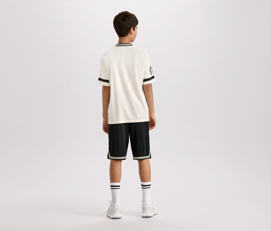Hátulról látható egy fiatalember, aki rövid ujjú, krémszínű inget visel, fekete csíkokkal a gallérján és az ujján, fekete rövidnadrágot fehér csíkokkal, valamint fehér zoknit és cipőt.