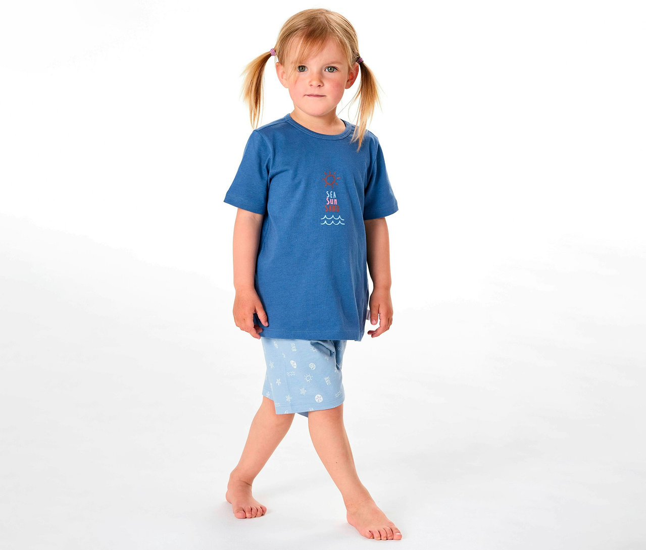 Egy kislány SCHIESSER rövidnadrágos gyerekpizsamát visel, kék felsővel és világoskék mintás rövidnadrággal.