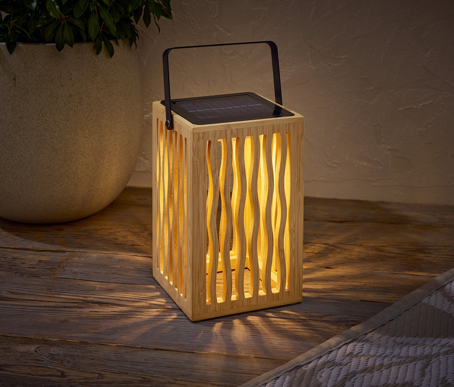 Egy fa napelemes lámpa fekete fogantyúval egy fa asztalon áll egy növényes cserép mellett.