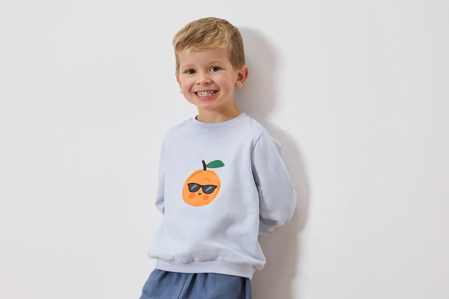 Lächelnder Junge trägt hellblaues Sweatshirt mit einer Orange mit Sonnenbrille darauf.