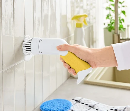 Egy kéz egy sárga nyelű, fehér fejű elektromos tisztítókefét tart, amivel csempét tisztít.