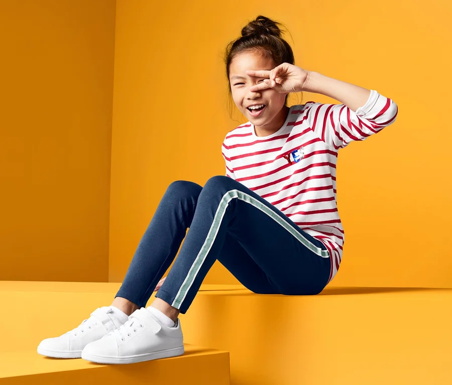 Egy lány ül egy sárga háttér előtt, és eltakarja az egyik szemét. 2 lány leggings szettben és egy csíkos pólót visel.