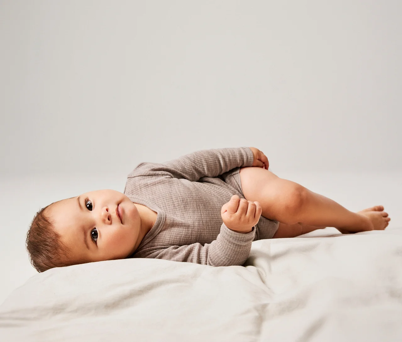 Baba fekszik a hátán egy világos lepedőn, bordázott, hosszú ujjú, taupe színű bodyt visel, és a kamerába néz.
