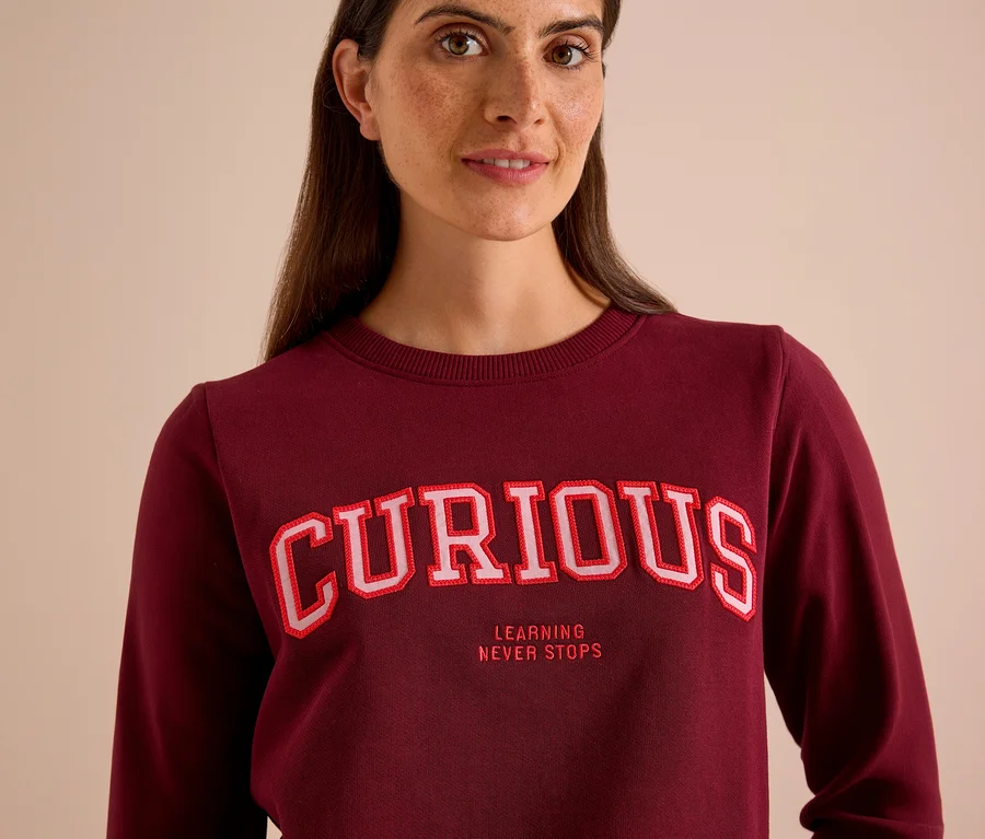 Nő visel egy bordó CECIL szabadidőfelsőt, rajta „CURIOUS” és „LEARNING NEVER STOPS” felirattal.