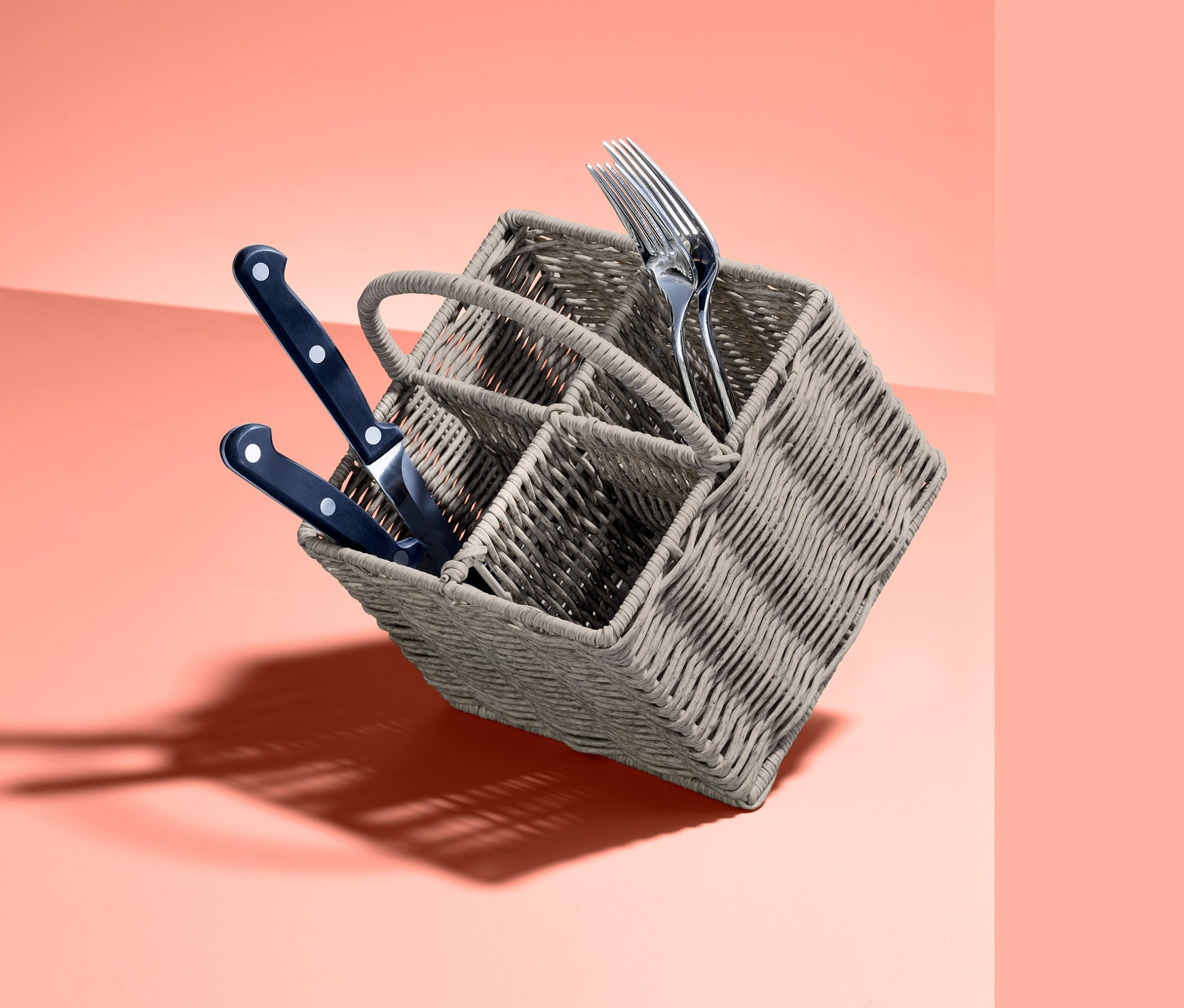 Szürke evőeszköztartó kosár késekkel és villákkal korall háttér előtt.