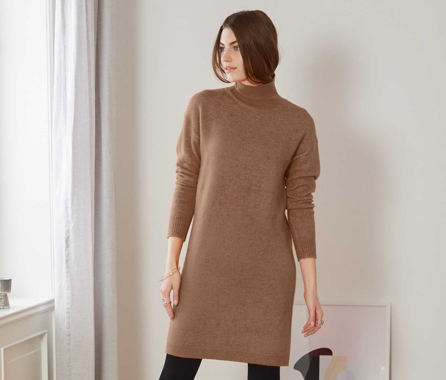 Nő pózol barna kötött ruhában, fekete leggingsben és csizmában.