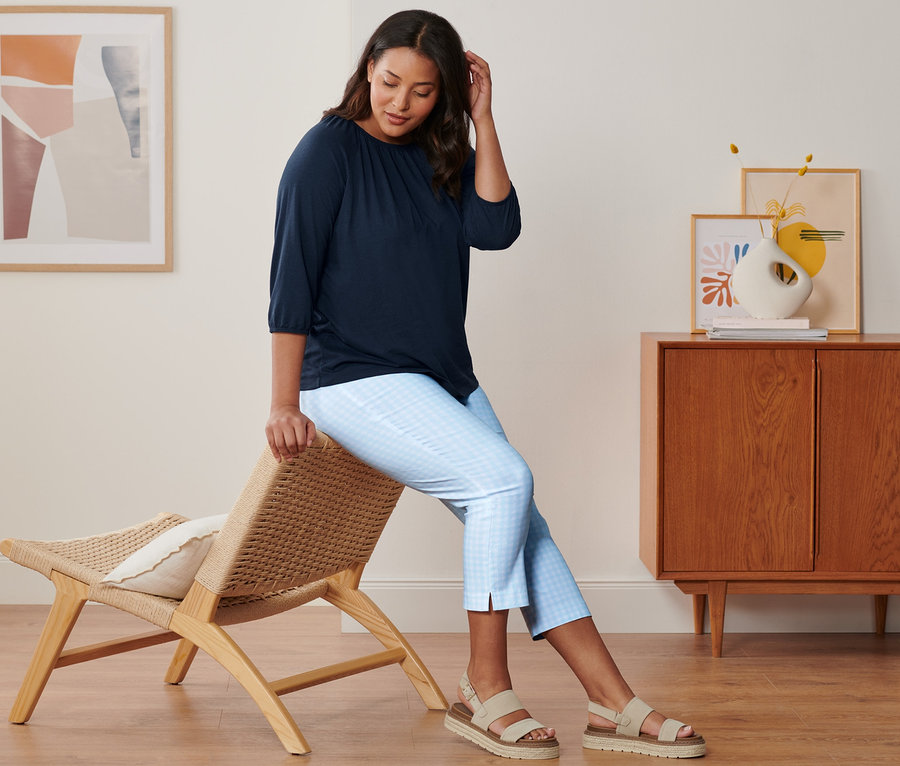 Nő ül egy széken, sötétkék 3/4-es ujjú felsőt, világoskék-fehér 7/8-os sztreccsnadrágot és bézs bőr szandált visel.