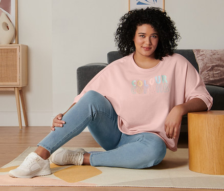 Egy nő ül a nappali padlóján. Rózsaszín, 3/4-es ujjú szabadidőfelsőt, treggingst és velúrbőr sneaker cipőt visel.
