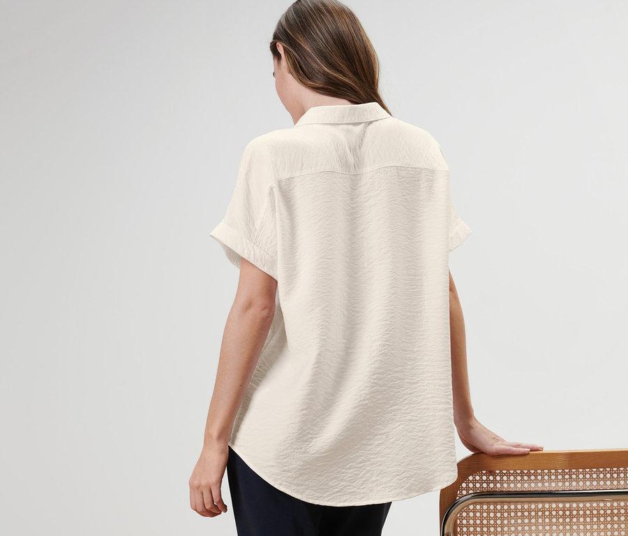 Egy nő háttal áll, blúzt visel és egy széknek támaszkodik.