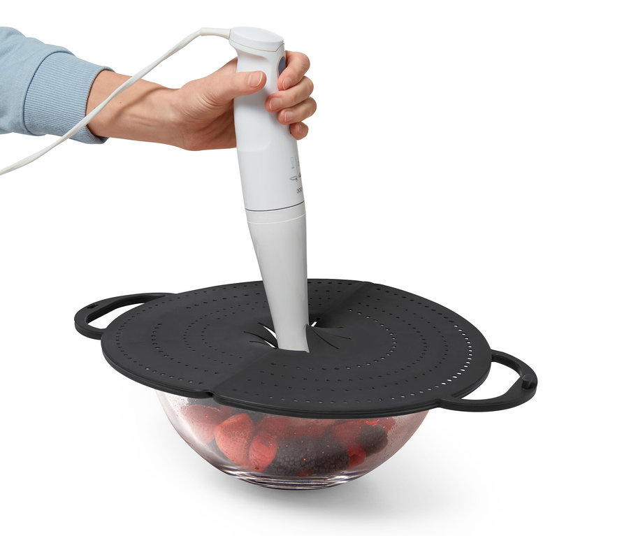 Egy kéz egy botmixert tart egy fekete, 4 az 1-ben konyhai mindenesen keresztül, ami egy üvegtál fölött van, tele gyümölcsökkel.