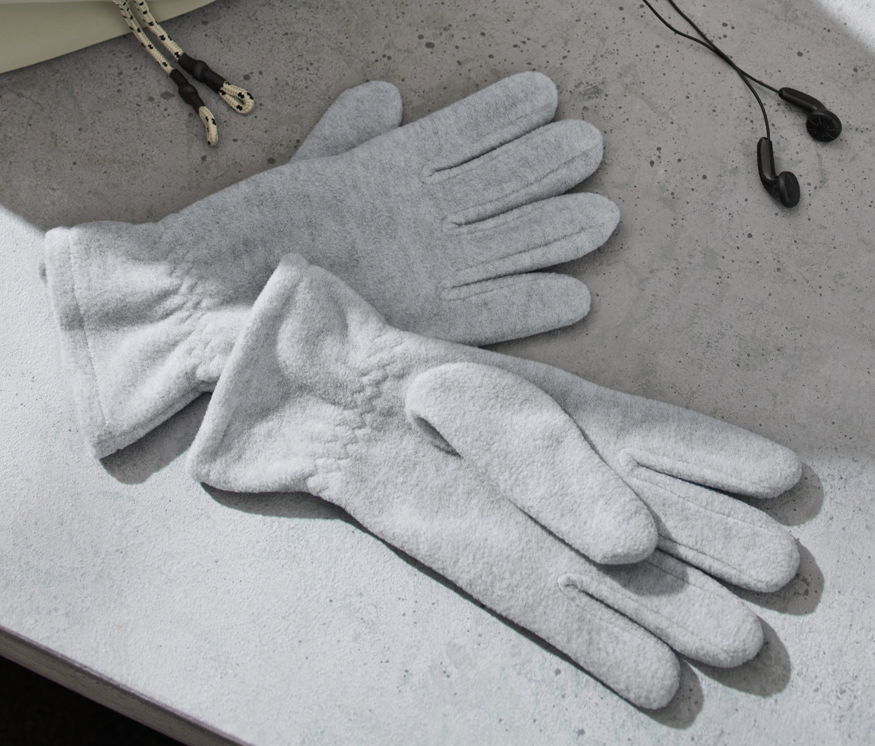 Két szürke polárkesztyű fekszik egy szürke felületen, fejhallgató és egy táska mellett.