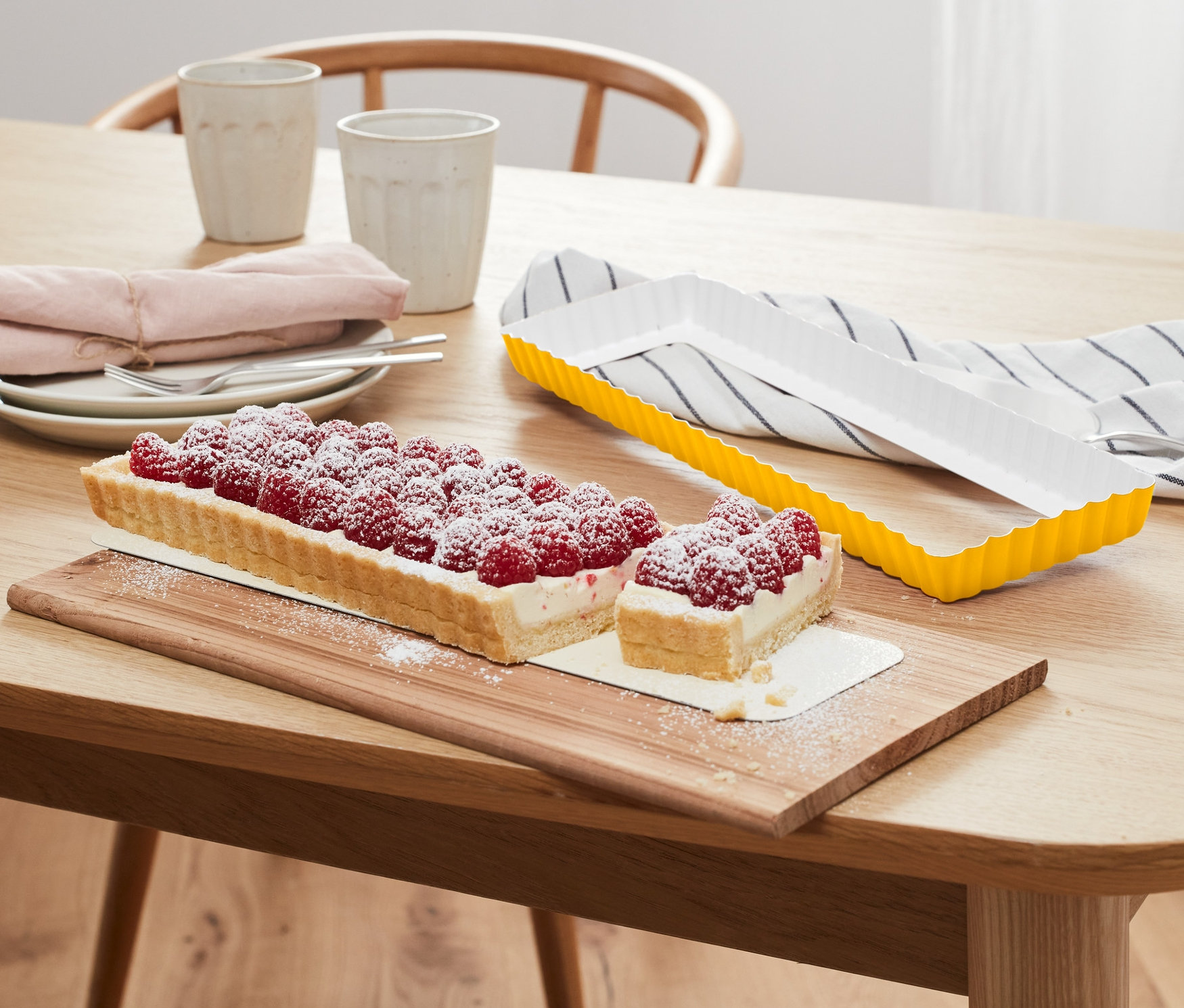 Téglalap alakú málnás torta fa deszkán, mellette egy sárga szögletes torta sütőforma.