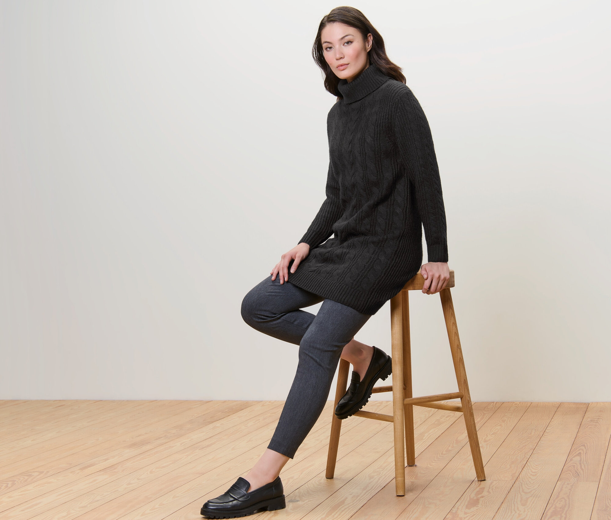 Egy nő ül egy széken, és egy hosszú, fekete, fonott mintás kötött pulóvert visel.