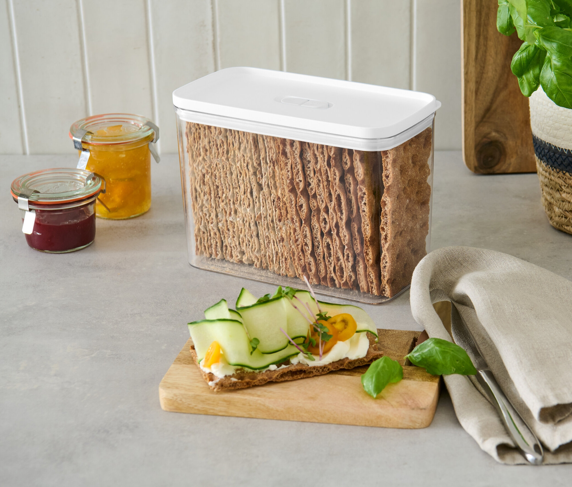 Egy fehér fedelű frissentartó doboz tele kenyérkekszekkel egy pulton áll lekváros üvegek mellett.