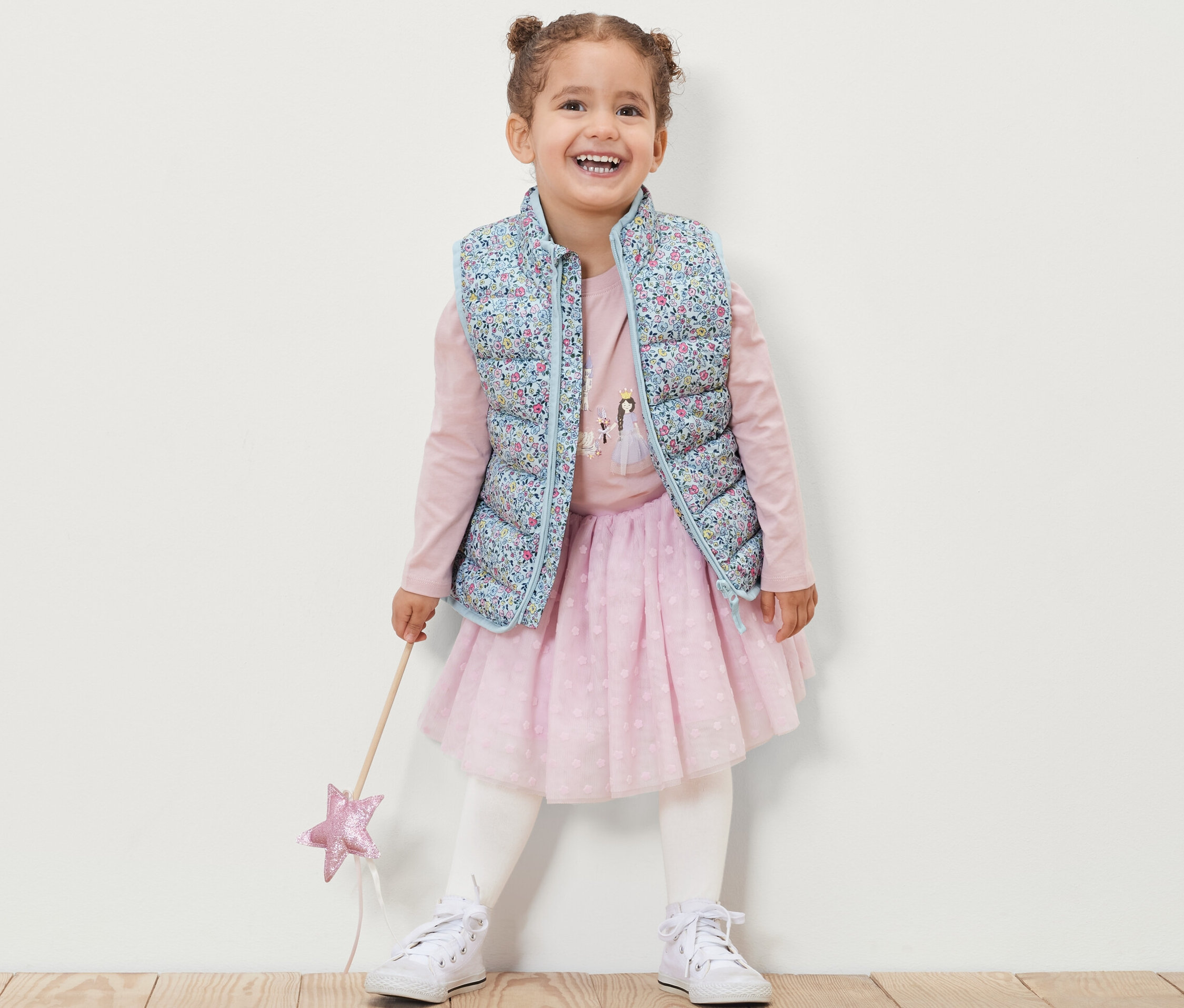 Mosolygó kislány virágos kislány steppelt mellényt, rózsaszín hosszú ujjú pólót, rózsaszín szoknyát és fehér tornacipőt visel, és egy varázspálcát tart.