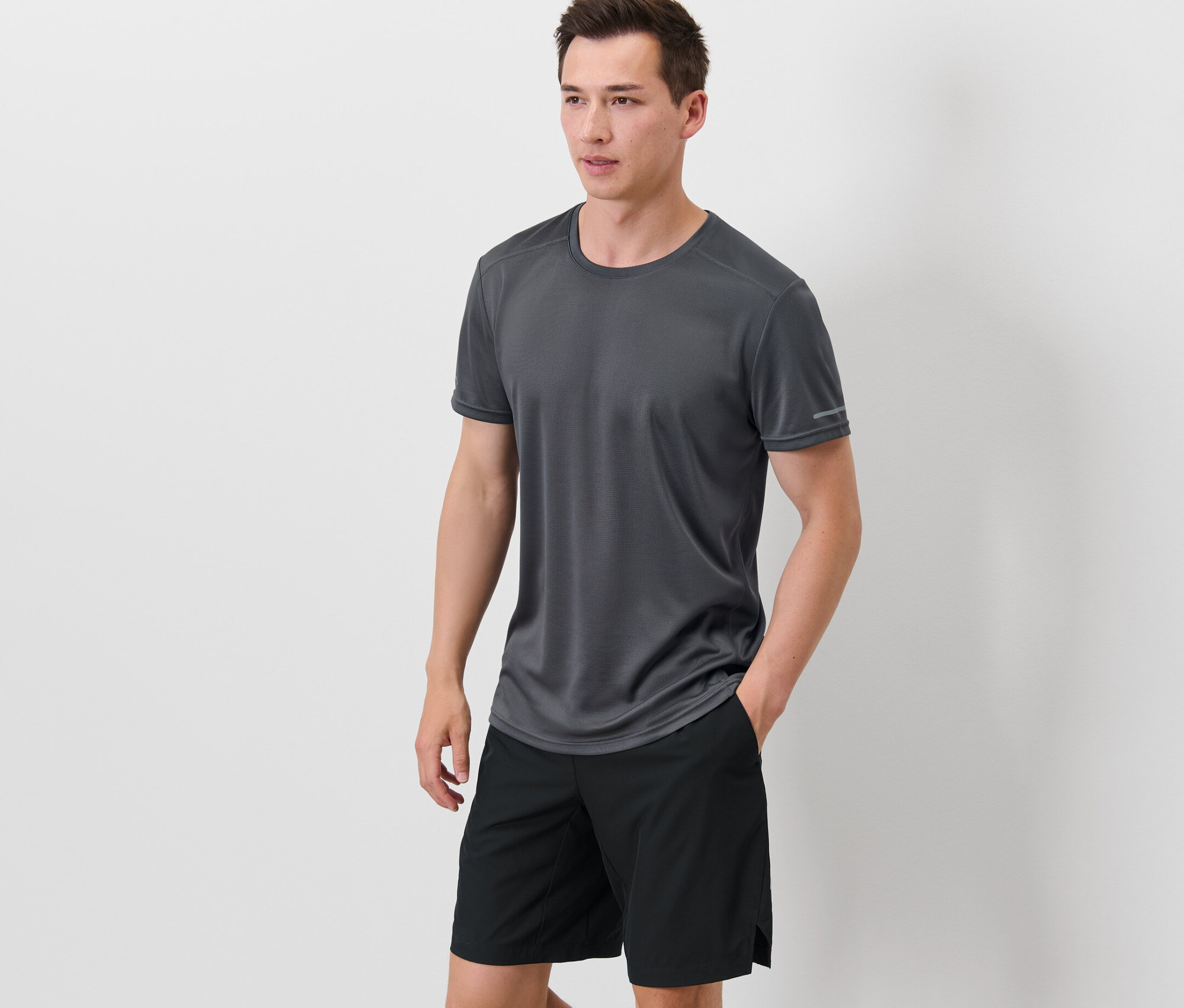 Egy férfi áll, kezével a fekete rövidnadrágja zsebében, szürke funkcionális felsőt visel.