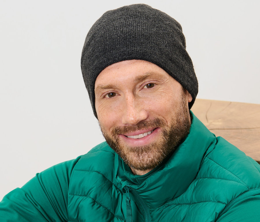 Szakállas férfi mosolyog, kötött sapkát és zöld mellényt visel.