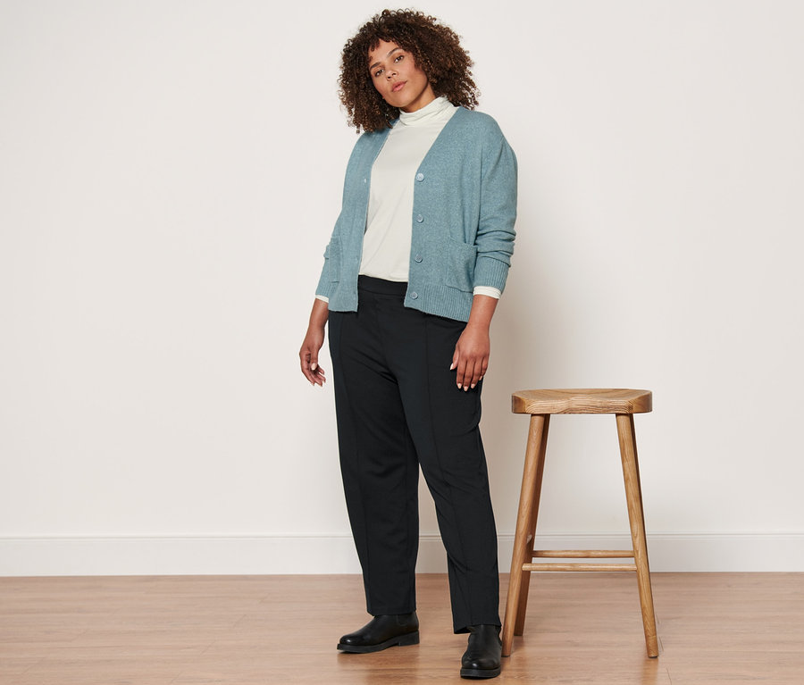 Egy nő egy fa zsámoly mellett áll, és gyapjú kötött kardigánt, egy krém garbót, egy fekete dzsörzénadrágot és egy bőrcsizmát visel.