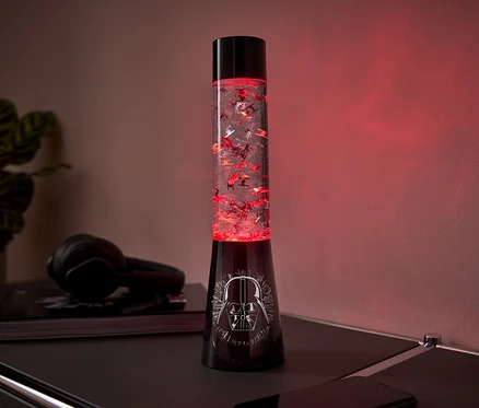 Egy fekete lávalámpa Darth Vader mintával az alján és piros csillámokkal a belsejében egy asztalon áll.